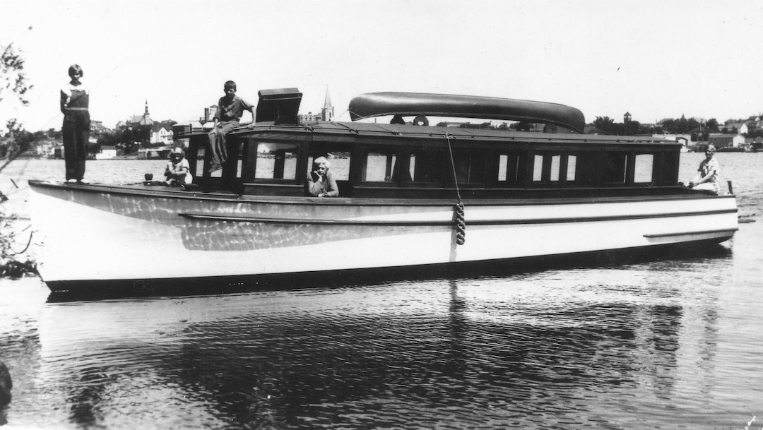 Black and white photograph. The Stone-built launch, Atus, with some of the members of the Stone family aboard. The boat is at Bush Island with the townscape in the background.