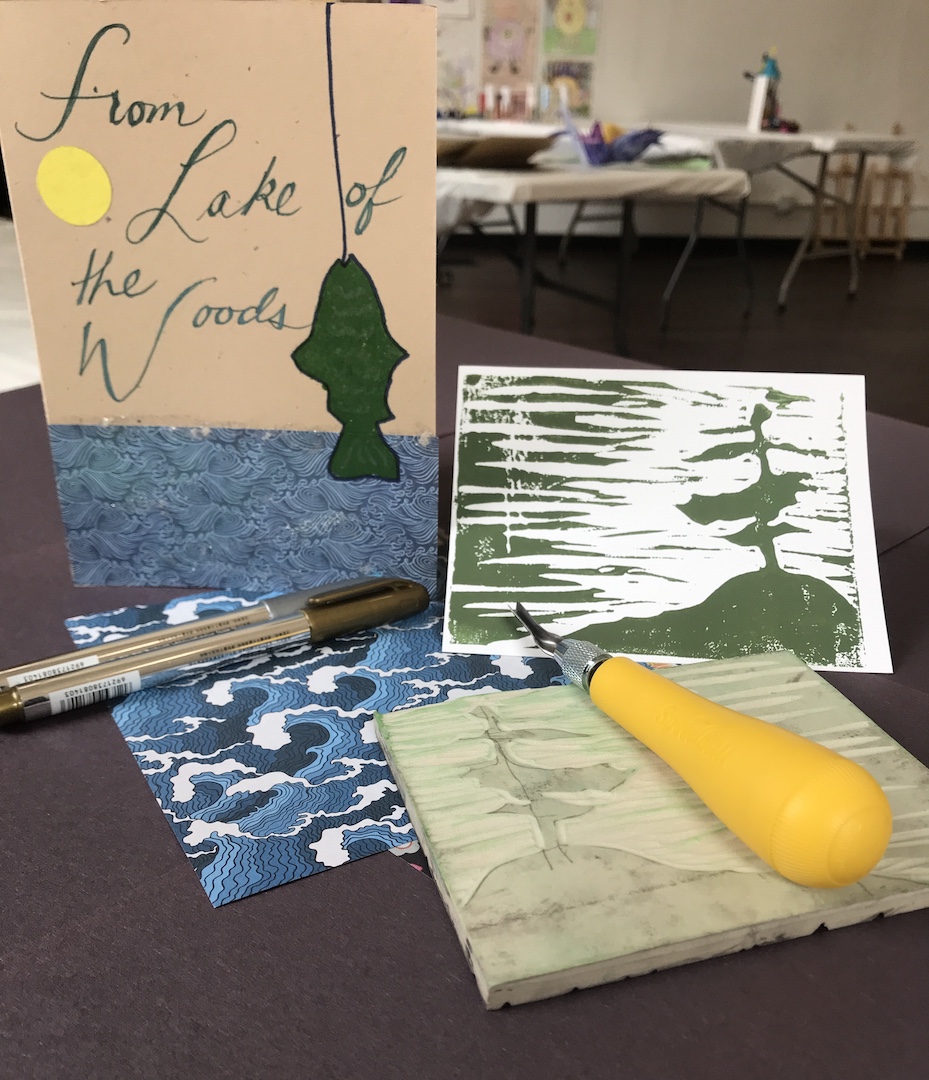 One card with a fix and water that says from Lake of the Woods with Love and another card with a lino block print of a tree, island and lake. A lino block, tool, origami paper and pens