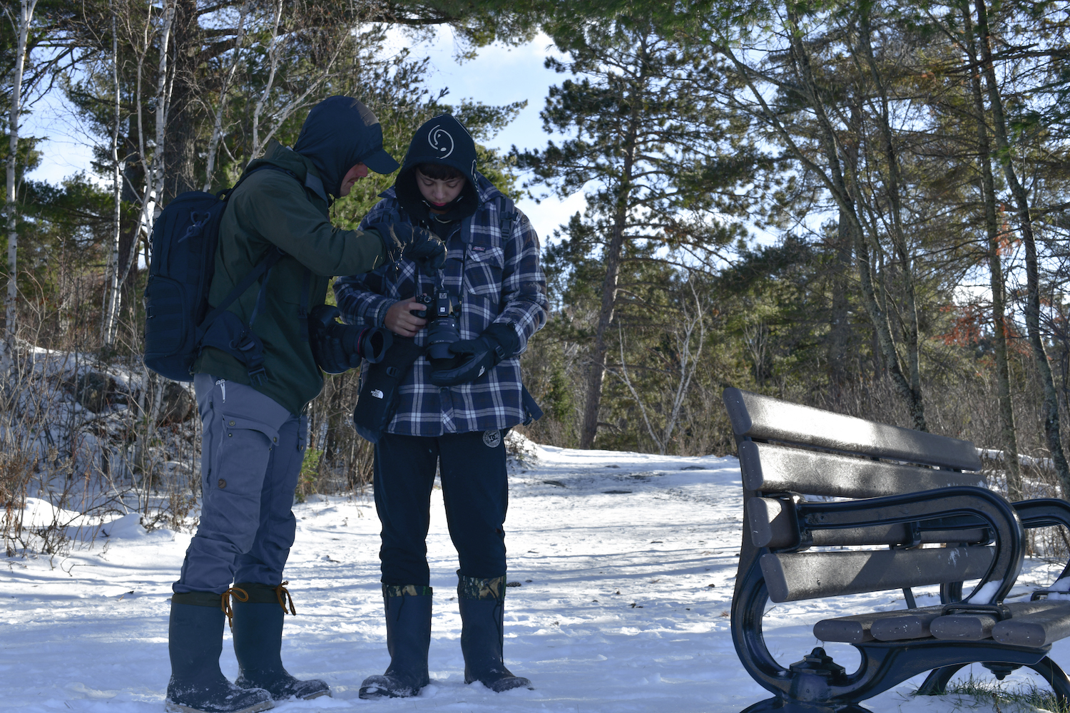 Two students from Beaver Brae Secondary School ACE program holding and looking at cameras outside in the winter during their the Intro to Digital Photography class.