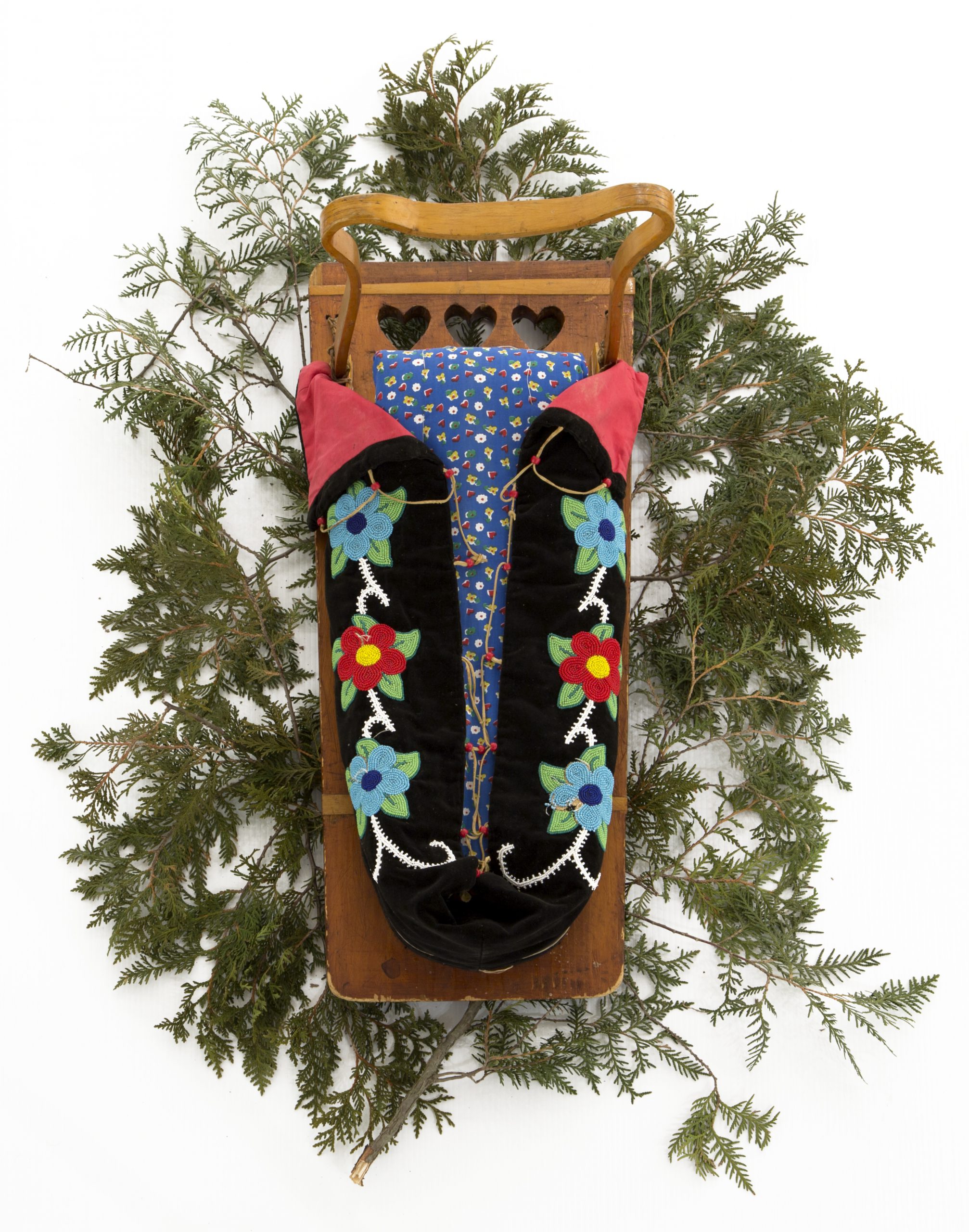 Photo of a tikinagaan (cradleboard) featuring traditional beading, placed on a backdrop of cedar branches.